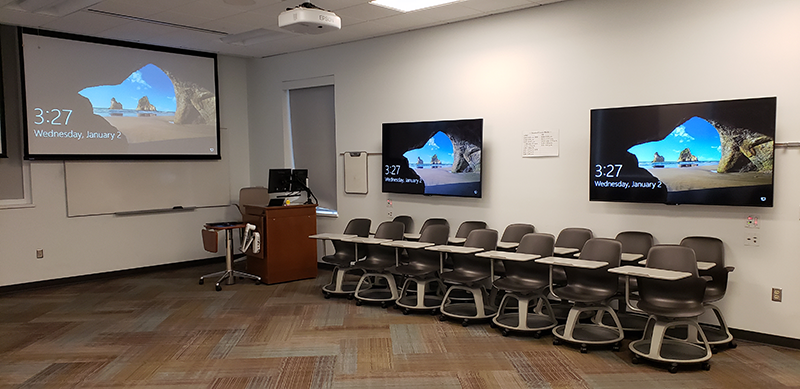 Moveable desks sit in rows in front of two wall-mounted display screens. A lectern with computer and projector screen sit to the left.